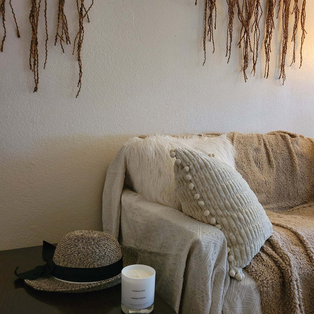 a taupe and cream sofa with fluffy neutral throw pillows, brown natural wall decor, a straw hat, and a good scents tumbler candler